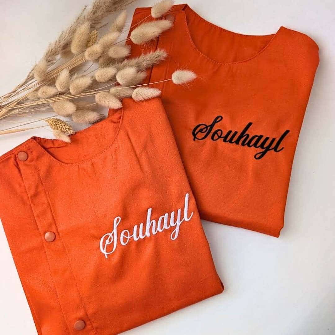 Tablier en tissus pour école maternelle couleur orange modèle sans manches pour garçon brodé du prénom Souhayl en noir